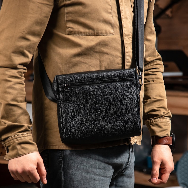 May include: A black pebbled leather crossbody bag featuring a rectangular shape, a top zipper closure, and a wide black adjustable fabric shoulder strap. The bag is worn across the torso, showcasing a textured surface and a minimalist design.