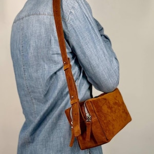 May include: A rectangular tobacco-brown suede crossbody bag featuring a long adjustable shoulder strap with metal hardware and a silver-toned zip closure. The bag has a soft, textured finish and is worn over a light blue denim button-down shirt.