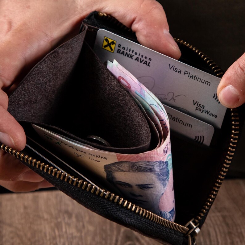 May include: A black leather zipped wallet containing a Raiffeisen Bank Aval Visa Platinum card and a Ukrainian banknote featuring a portrait. The wallet has a dark brown interior lining and a gold-toned metal zip along the top edge.