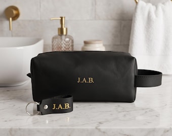 May include: A black leather wash bag with a side handle and a matching black leather keyring, both featuring the gold embossed initials J.A.B. The rectangular bag has a zip closure and a smooth finish, presented as a coordinated set.