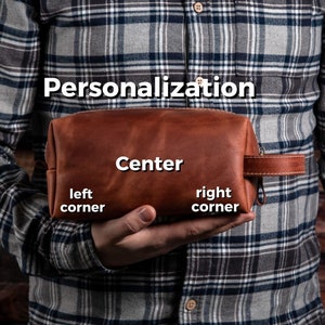 May include: A brown leather toiletry bag with a side handle and a zip closure. The bag features text overlays indicating personalisation options, including the words Personalisation, Center, left corner, and right corner, displayed on the surface.