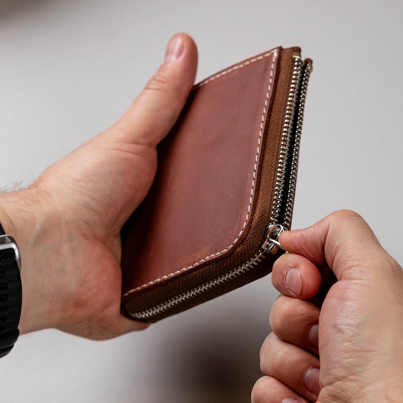 May include: A brown leather wallet with a smooth finish and visible white stitching along the edges. The wallet features a silver-toned metal zip that runs along the side, allowing for secure closure of the main compartment.