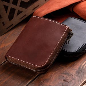 May include: A brown leather wallet with a wrap-around zip closure and visible white stitching along the edges. The wallet features a smooth, matte finish and a metal zip pull. A second black leather wallet is partially visible behind it.