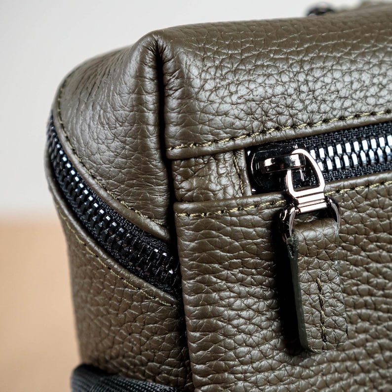 May include: An olive green bag with a textured, pebbled surface. The bag features black zippers with metallic silver pulls and a matching leather pull tab. The construction includes visible stitching along the seams and a structured, rectangular corner.