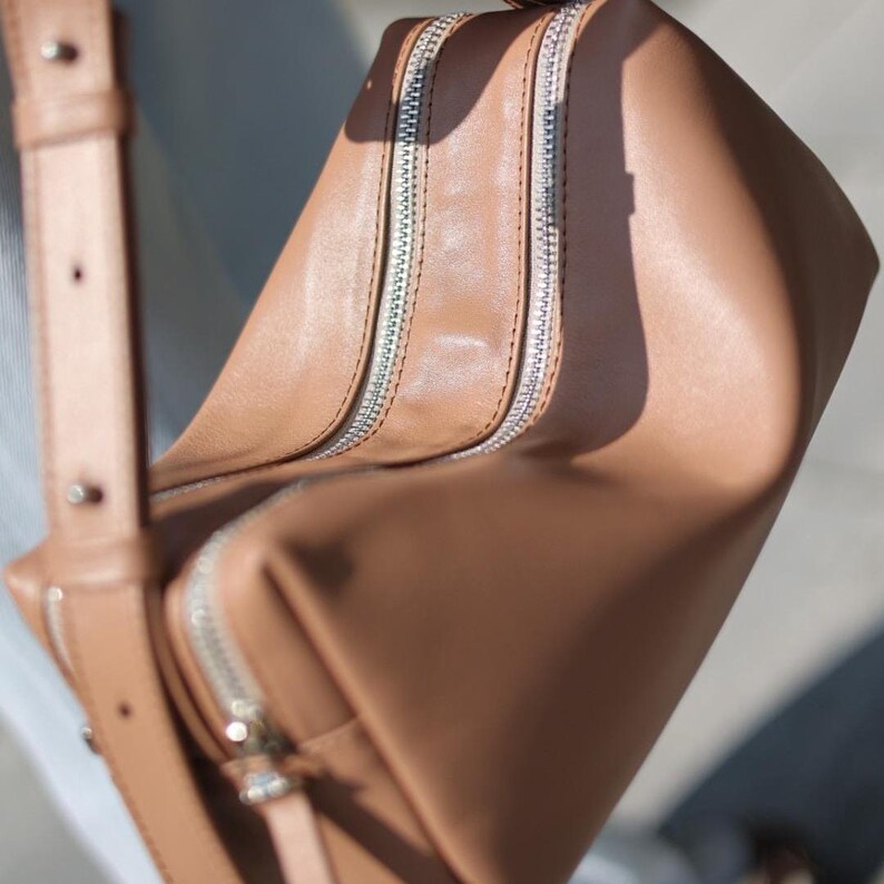 Pu&ograve; includere: Una borsa a tracolla color cuoio con superficie liscia, due cerniere metalliche argentate ben visibili sulla parte superiore e una tracolla regolabile in tinta con borchie metalliche argentate per la regolazione.