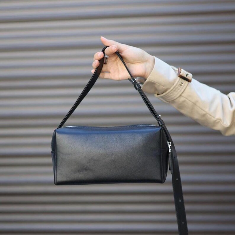 Pu&ograve; includere: Una borsa a mano rettangolare nera con finitura liscia, dotata di chiusura superiore con cerniera e una sottile tracolla nera regolabile con fibbia in metallo color argento. La borsa presenta una forma strutturata e un esterno nero uniforme.