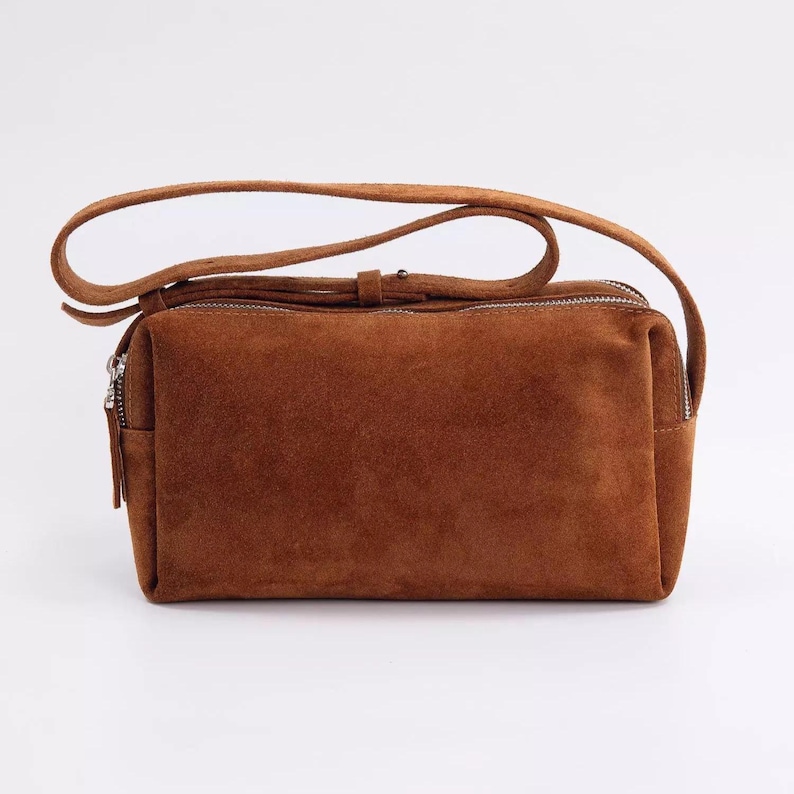 May include: A rectangular brown suede shoulder bag featuring a smooth texture, a single adjustable strap with a metal buckle, and a silver-toned zip closure along the top edge. The bag has a structured, boxy shape with clean, finished seams.
