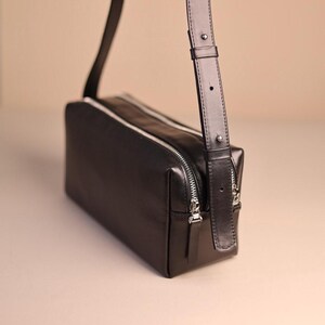 Pu&ograve; includere: Una borsa a tracolla rettangolare nera con finitura liscia, dotata di chiusura superiore con zip, dettagli argentati e una tracolla regolabile con un elemento metallico. La borsa presenta una silhouette strutturata e squadrata.