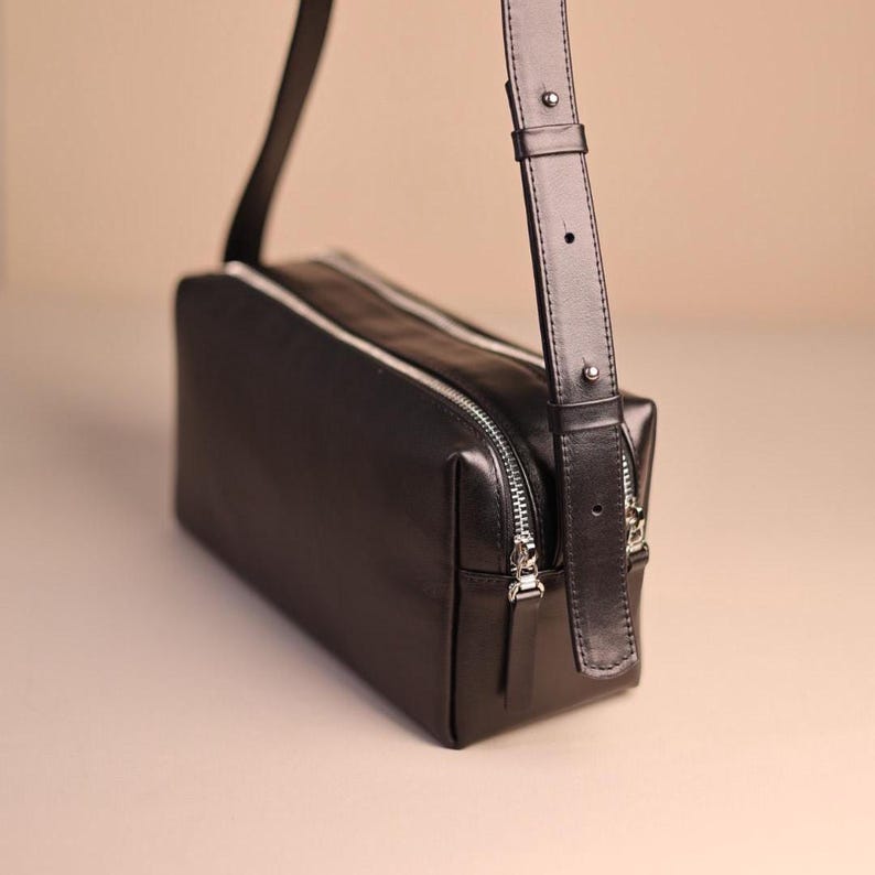 Pu&ograve; includere: Una borsa a tracolla rettangolare nera con finitura liscia, dotata di chiusura superiore con zip, dettagli argentati e una tracolla regolabile con un elemento metallico. La borsa presenta una silhouette strutturata e squadrata.