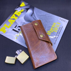 Puede incluir: Una cartera de cuero marr&oacute;n con cierre de solapa asegurado por dos broches de metal color lat&oacute;n, con costuras blancas visibles a lo largo de los bordes, acompa&ntilde;ada de un encendedor de metal cepillado color lat&oacute;n con la tapa abierta.
