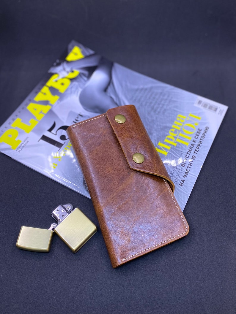 Puede incluir: Una cartera de cuero marr&oacute;n con cierre de solapa asegurado por dos broches de metal color lat&oacute;n, con costuras blancas visibles a lo largo de los bordes, acompa&ntilde;ada de un encendedor de metal cepillado color lat&oacute;n con la tapa abierta.