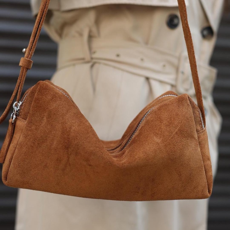 Pu&ograve; includere: Una borsa a tracolla in camoscio marrone dalla silhouette rettangolare, dotata di chiusura superiore con zip, dettagli color argento e una tracolla regolabile in camoscio coordinato. La borsa presenta una finitura morbida.