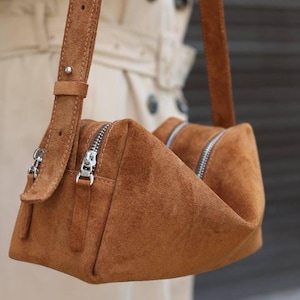May include: A tan suede shoulder bag featuring a structured, triangular silhouette with two silver-toned metal zips along the top. The bag includes a matching adjustable suede shoulder strap attached with a silver metal stud and buckle hardware.