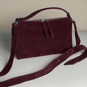 May include: A deep burgundy suede handbag featuring a rectangular silhouette, a top handle, and a long adjustable shoulder strap. The bag includes a silver-toned metal zipper closure with two matching suede pull tabs and silver hardware accents.