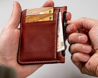 May include: A brown leather wallet featuring white contrast stitching, multiple card slots containing two gold-coloured credit cards, and a side zippered compartment holding a Ukrainian banknote with visible text reading UKRAЇNA.