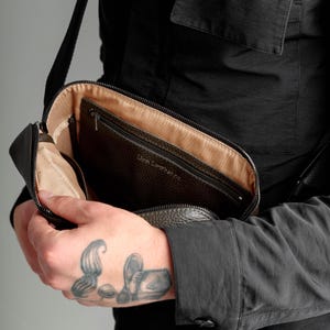 May include: A dark brown pebbled leather crossbody bag with a zipper closure and a beige fabric interior lining. The interior features a zippered pocket with the embossed text Lion Leather Co. The bag has a black adjustable shoulder strap.