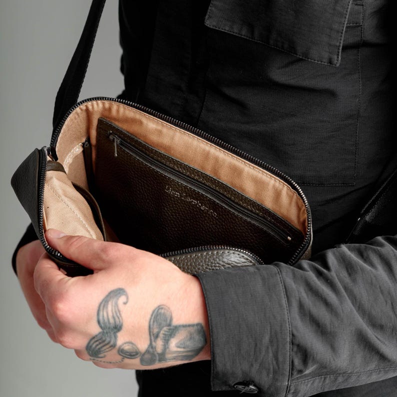 May include: A dark brown pebbled leather crossbody bag with a zipper closure and a beige fabric interior lining. The interior features a zippered pocket with the embossed text Lion Leather Co. The bag has a black adjustable shoulder strap.