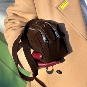Pu&ograve; includere: Una borsa a mano in camoscio marrone scuro di forma rettangolare, dotata di tracolla regolabile in tinta e due cerniere in metallo argentato sulla parte superiore. La borsa &egrave; tenuta contro un cappotto color cammello, con le cerniere ben visibili.