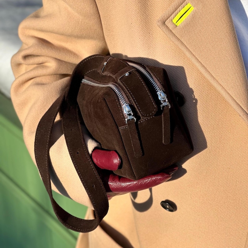 Pu&ograve; includere: Una borsa a mano in camoscio marrone scuro di forma rettangolare, dotata di tracolla regolabile in tinta e due cerniere in metallo argentato sulla parte superiore. La borsa &egrave; tenuta contro un cappotto color cammello, con le cerniere ben visibili.