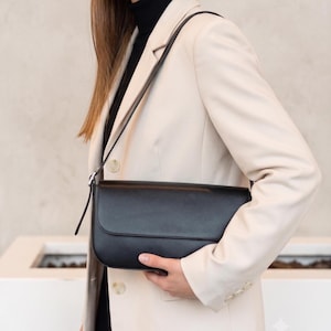 Può includere: Una borsa a tracolla in pelle nera liscia con bordo inferiore arrotondato e chiusura a patta. La borsa presenta una tracolla sottile e regolabile con una fibbia in metallo argentato, indossata sopra un blazer beige chiaro e un dolcevita.