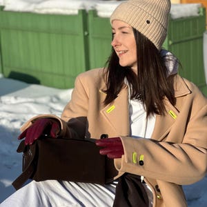 Pu&ograve; includere: Un berretto in maglia a coste beige, un cappotto in lana color cammello con dettagli asola giallo neon, una felpa bianca con cappuccio e guanti in pelle rosso scuro. Una borsa rettangolare marrone scuro con cerniera argentata e cinturino.