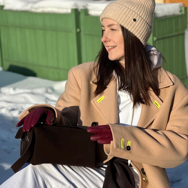Pu&ograve; includere: Un berretto in maglia a coste beige, un cappotto in lana color cammello con dettagli asola giallo neon, una felpa bianca con cappuccio e guanti in pelle rosso scuro. Una borsa rettangolare marrone scuro con cerniera argentata e cinturino.