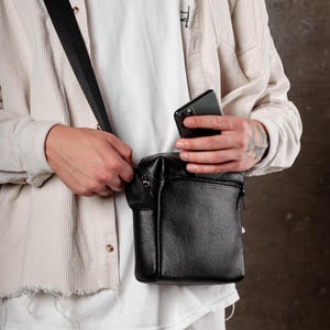 May include: A black pebbled leather crossbody bag with a rectangular shape, a top zipper closure, and an adjustable black fabric shoulder strap. The bag features a front zippered pocket and is shown with a dark gray smartphone being placed inside.