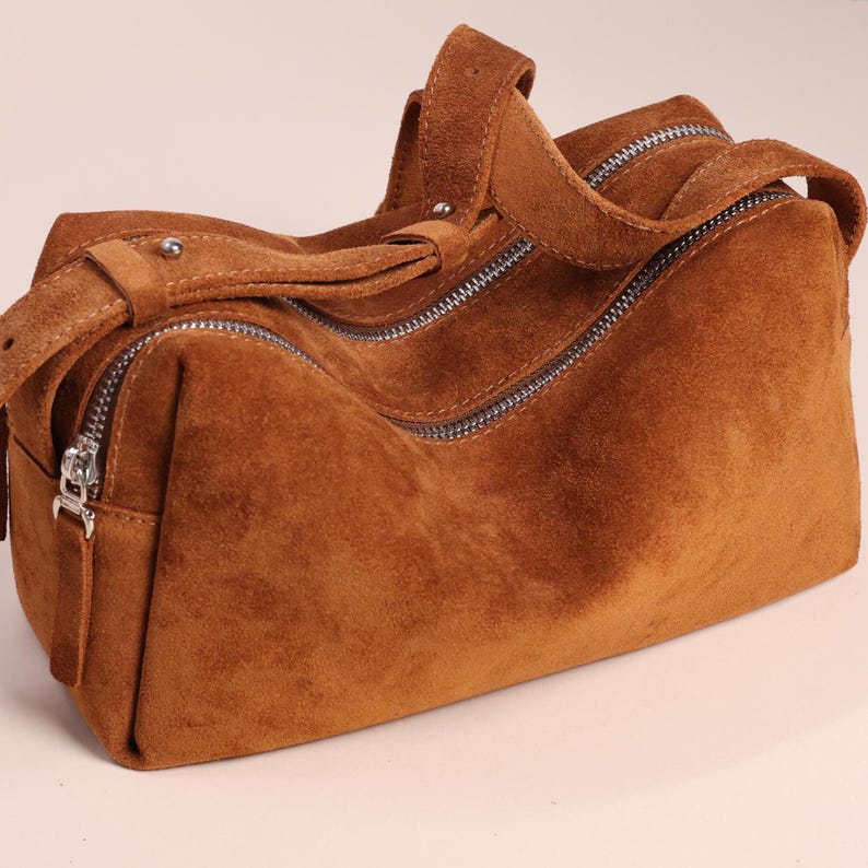 Pu&ograve; includere: Una borsa a mano rettangolare realizzata in materiale scamosciato marrone. La borsa presenta un manico superiore con un rivetto in metallo argentato, uno scomparto principale chiuso da una cerniera color argento e una base strutturata.