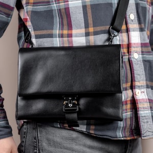 May include: Black crossbody bag with a flap closure and a metal clasp. The bag has a black adjustable strap and is worn over a plaid shirt and jeans. The bag appears to be made of leather.