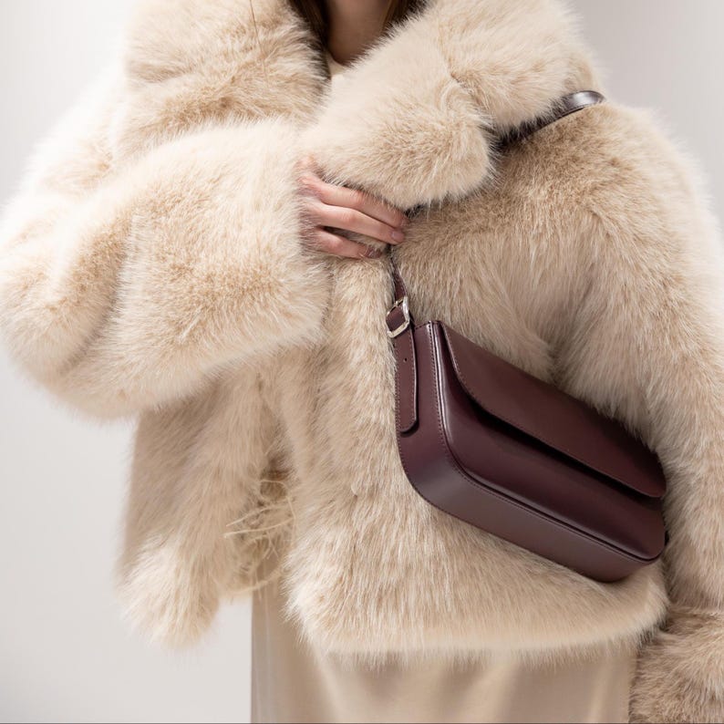 May include: A dark burgundy leather shoulder bag with a flap closure and a silver-toned metal buckle on the strap, worn over a beige faux fur jacket with long, thick pile texture and a high collar, paired with a plain beige garment underneath.