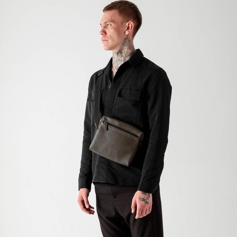 May include: A dark olive green rectangular crossbody bag with a top zipper closure and an adjustable shoulder strap, worn over a black long-sleeved button-down shirt and dark trousers. The bag features a textured finish and a slim profile.