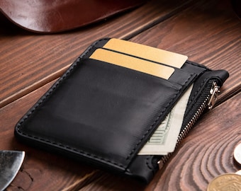 May include: A black leather card holder wallet featuring a side zip compartment, visible stitching along the edges, and two gold-coloured cards inserted into the top slots with a portion of a paper banknote visible inside.