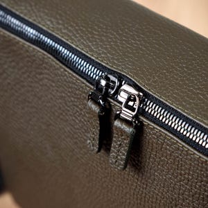 May include: An olive green bag with a textured, pebbled surface featuring a black zipper closure. The zipper has two metallic silver pulls, each attached to a matching olive green leather tab with visible stitching along the edges of the tabs.
