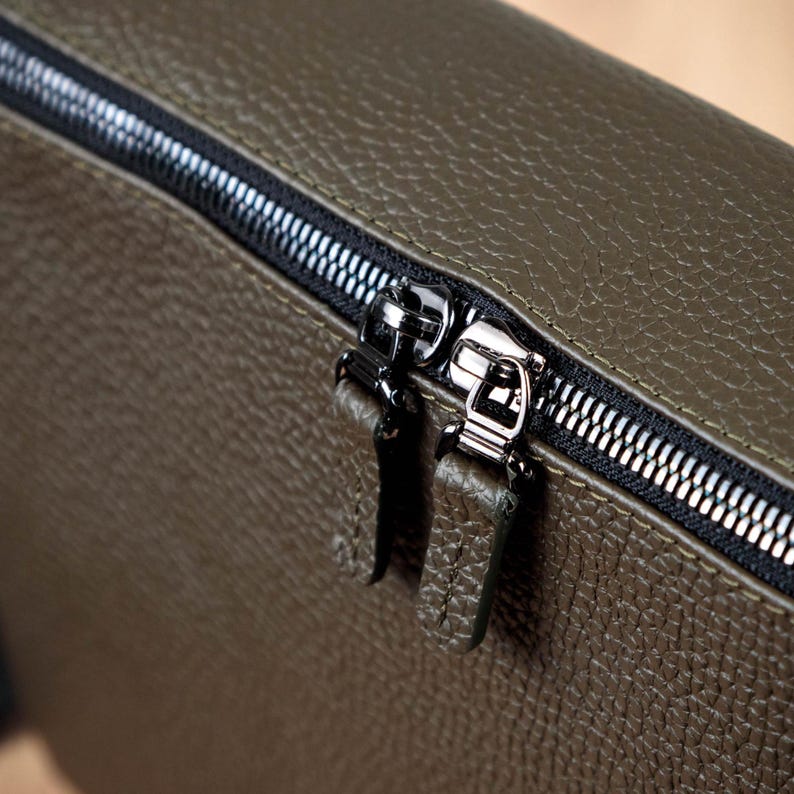 May include: An olive green bag with a textured, pebbled surface featuring a black zipper closure. The zipper has two metallic silver pulls, each attached to a matching olive green leather tab with visible stitching along the edges of the tabs.