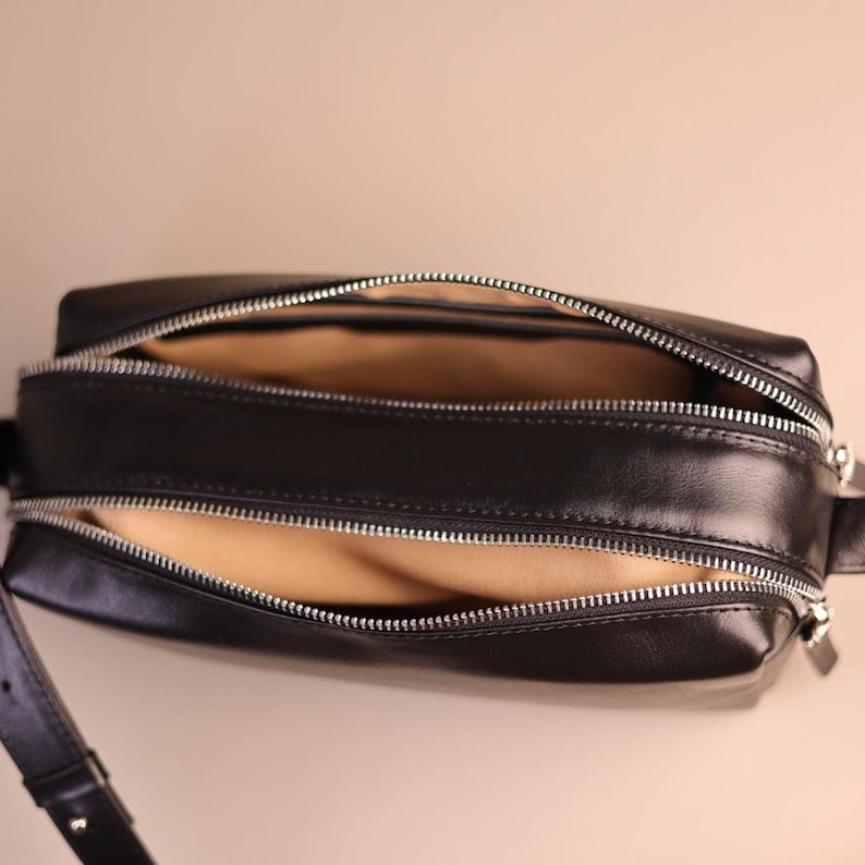 Pu&ograve; includere: Una borsa a tracolla rettangolare nera con due scomparti separati dotati di cerniera. La borsa presenta cerniere in metallo color argento e una finitura esterna liscia. L'interno di entrambi gli scomparti &egrave; foderato con un tessuto color marrone chiaro.