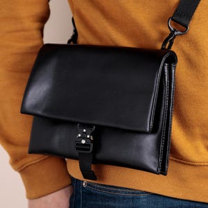 Puede incluir: Un bolso bandolera negro con cierre de solapa y hebilla metálica. El bolso tiene una correa negra ajustable y está hecho de un material liso. El bolso se lleva sobre un jersey color mostaza y unos vaqueros azules.