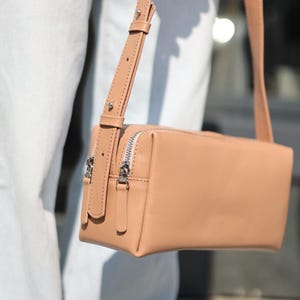 Pu&ograve; includere: Una borsa a tracolla rettangolare color beige con finitura liscia, dotata di una lunga tracolla regolabile con dettagli in metallo argentato e due chiusure a zip superiori con tiretti in pelle beige coordinati.