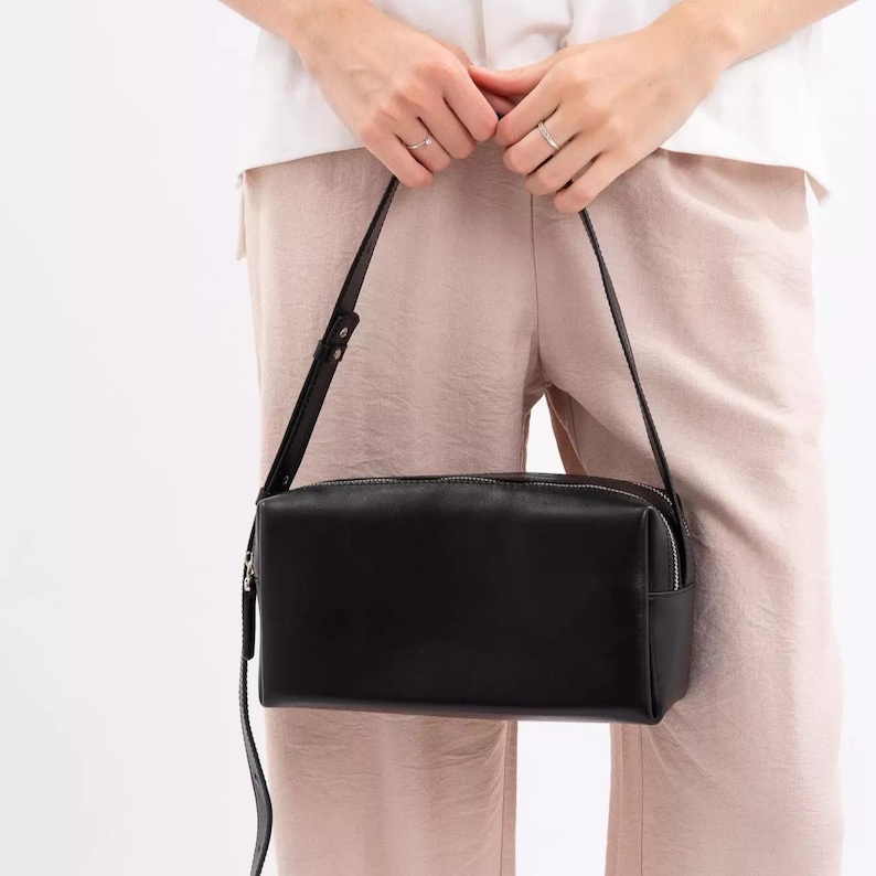 Pu&ograve; includere: Una borsa a tracolla rettangolare in pelle nera con finitura liscia, dotata di chiusura superiore con cerniera e una tracolla sottile regolabile. La borsa presenta una silhouette strutturata e squadrata con linee pulite e dettagli in metallo argentato.
