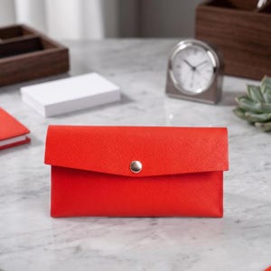May include: A rectangular red wallet with a textured surface and a single silver-toned snap button closure. The wallet features a flap design and is constructed from a durable material with a cross-hatched pattern visible on the exterior surface.
