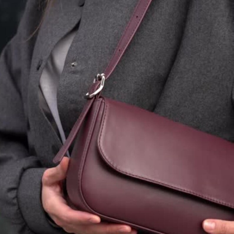 May include: A deep burgundy shoulder bag with a smooth, matte finish and a rounded flap closure. The bag features a matching adjustable strap attached with a silver-toned metal buckle hardware, providing a clean and structured appearance.