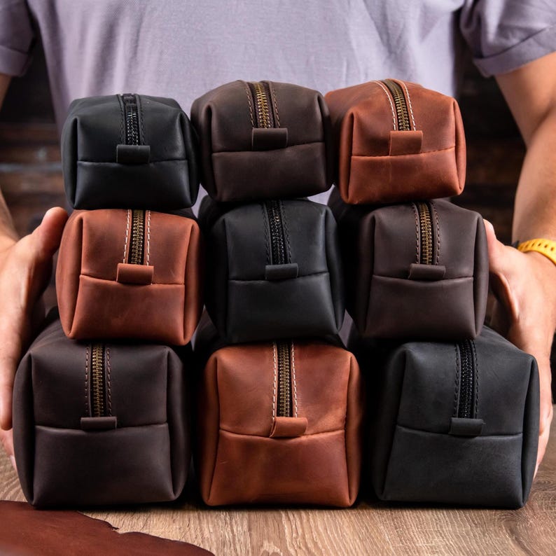 May include: A collection of rectangular leather pouches in various shades of black, dark brown, and tan. Each pouch features a central zip closure with a matching leather pull tab and visible stitching along the top seam of the bag.