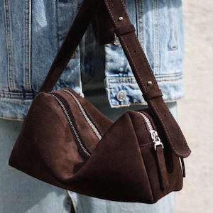 Pu&ograve; includere: Una borsa a tracolla in camoscio marrone scuro dalla forma strutturata e allungata. La borsa presenta una chiusura con cerniera in metallo color argento lungo la parte superiore e una tracolla in camoscio regolabile con un dettaglio a borchia argentata.