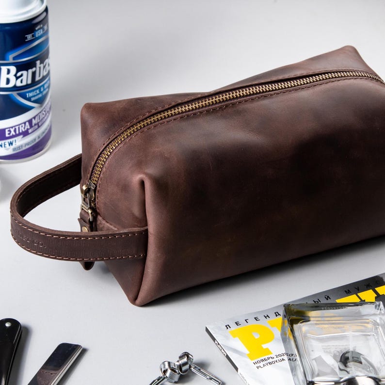 May include: A dark brown leather wash bag featuring a rectangular shape, a sturdy side handle, and a metallic zip closure. The bag has a smooth, matte finish and is constructed with visible stitching along the handle and main body seams.