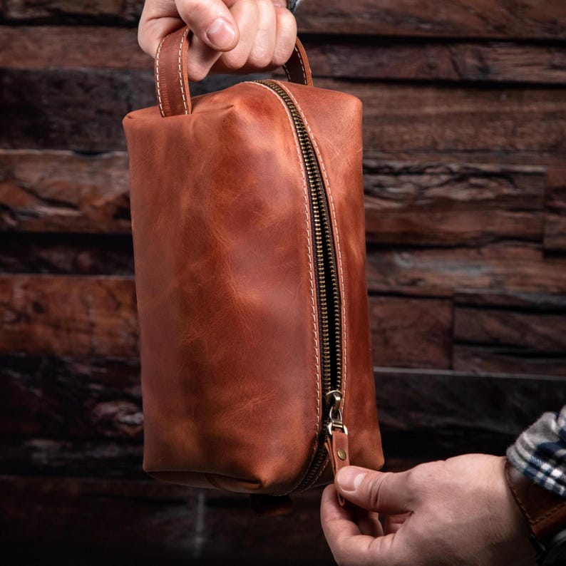 May include: A rectangular cognac-coloured leather toiletry bag featuring a top handle, a vertical brass-toned metal zip closure, and visible light-coloured stitching along the edges. The leather has a smooth, matte finish with natural variations.