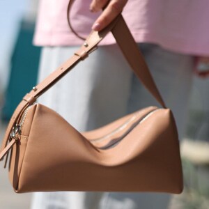 May include: A tan shoulder bag with a smooth, matte finish and a structured, rectangular silhouette. The bag features a single adjustable strap with silver-toned metal hardware and a top zip closure that runs across the length of the bag.