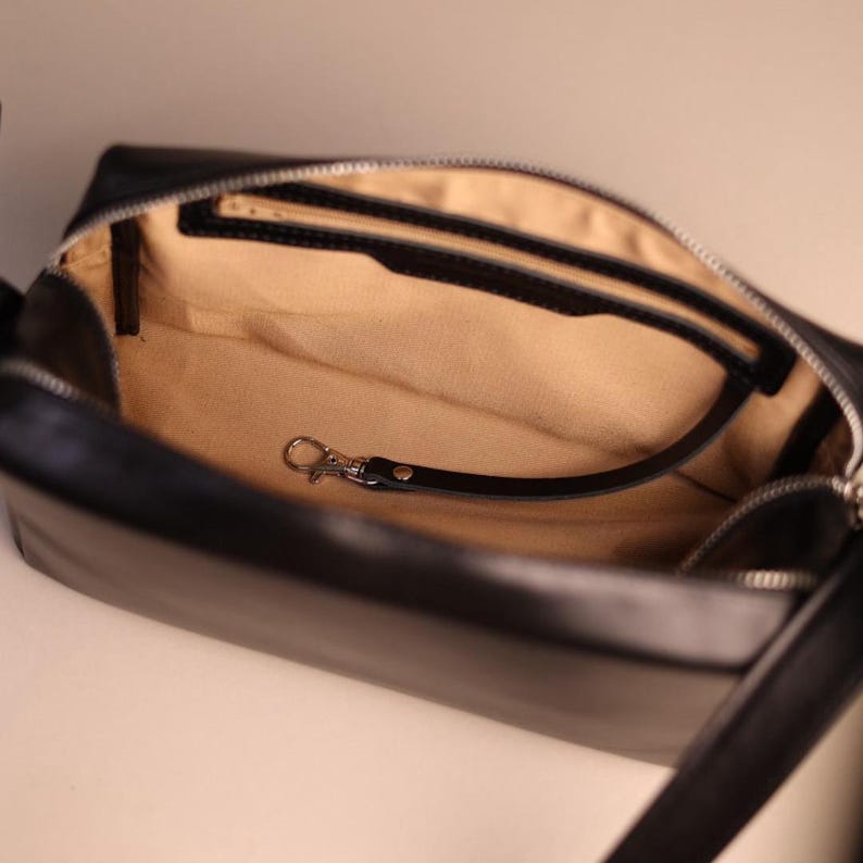 Pu&ograve; includere: Una borsa rettangolare nera con chiusura a zip argentata e fodera interna beige. L'interno presenta una tasca nera con zip e un cinturino in pelle nera con un moschettone in metallo argentato fissato sul lato.