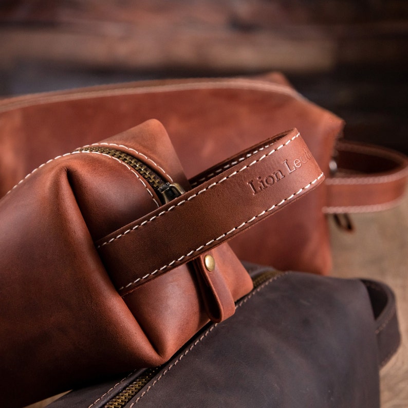 May include: A brown leather wash bag featuring a side handle with the embossed text Lion Leather, white contrast stitching, and a brass-toned metal zip. The bag has a structured rectangular shape with a smooth matte finish.