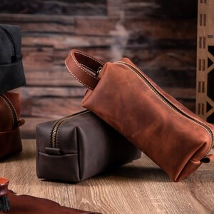 May include: A rectangular brown leather wash bag featuring a top handle with white stitching and a brass-toned metal zip closure. The bag has a smooth matte finish and a structured shape, positioned alongside other similar leather pouches.