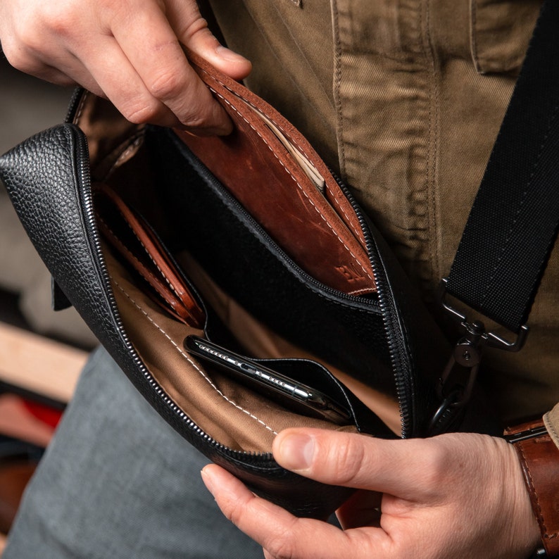 May include: A black pebbled leather crossbody bag with a zippered main compartment. The interior features a tan lining, a dedicated pocket containing a smartphone, and a separate brown leather sleeve insert with visible stitching and embossed text.