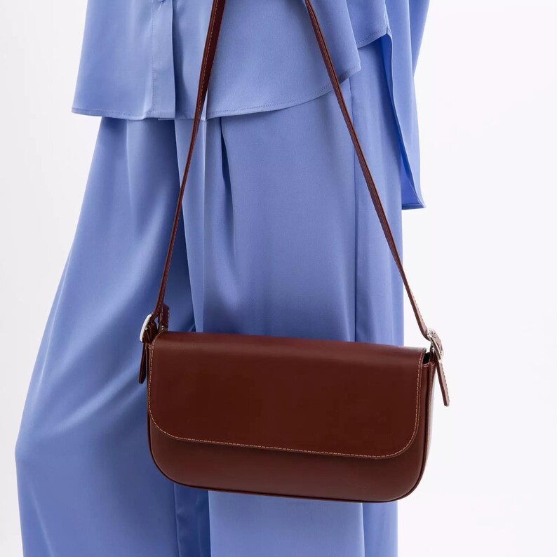 May include: A dark brown rectangular shoulder bag with a smooth finish and a flap closure. The bag features a thin, adjustable shoulder strap attached with silver-toned metal buckles on each side, providing a clean and structured appearance.